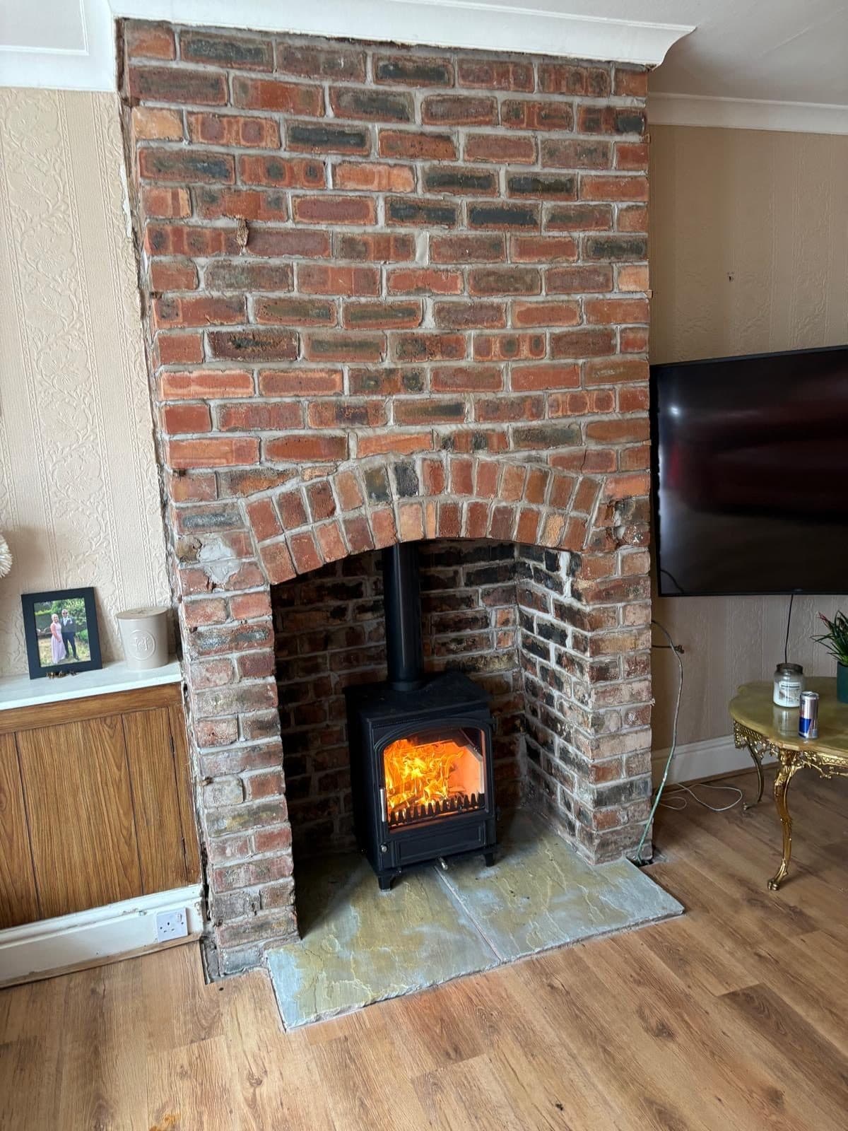 The same brick chimney breast with a small black stove installed and alight on a slate hearth