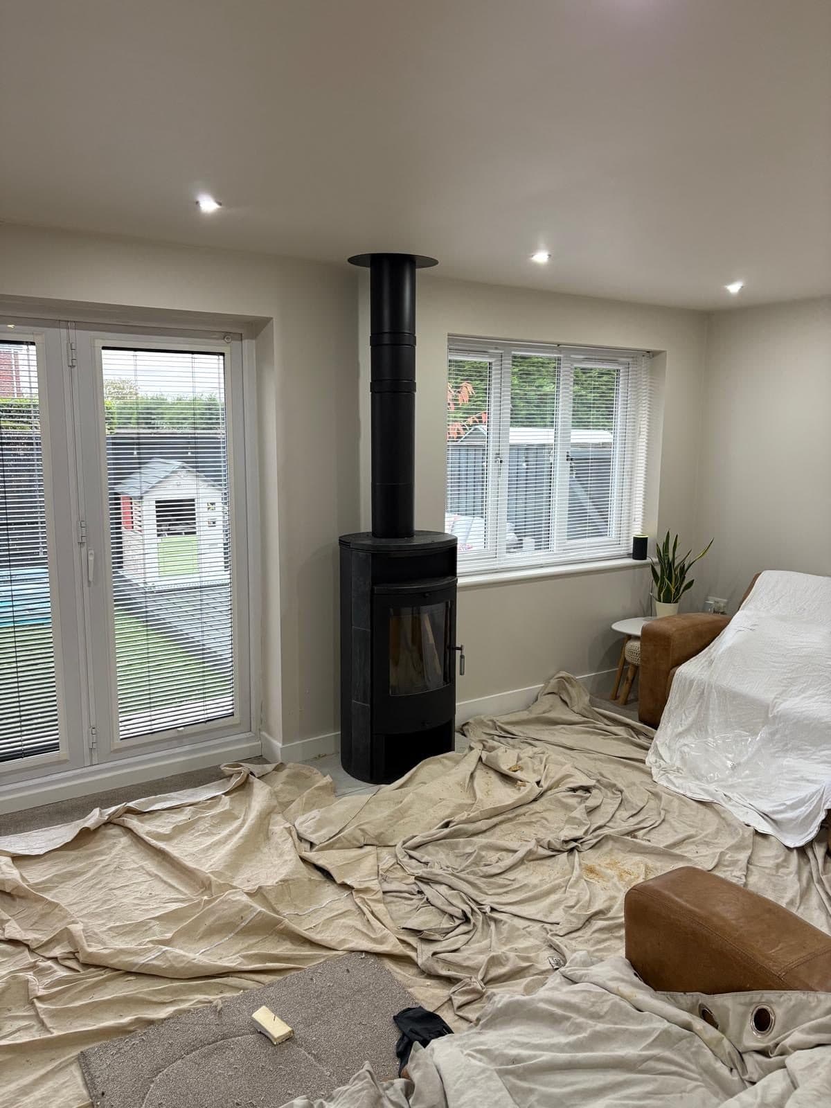 Lounge under dust sheets with a black cylindrical stove and twin-wall flue newly positioned, furniture covered