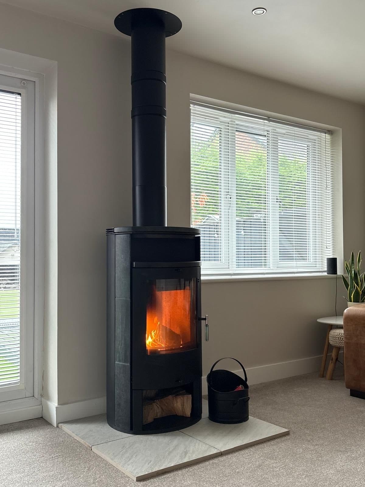 Tall black contemporary wood burner alight on a pale stone hearth, twin-wall flue running to the ceiling, log basket alongside