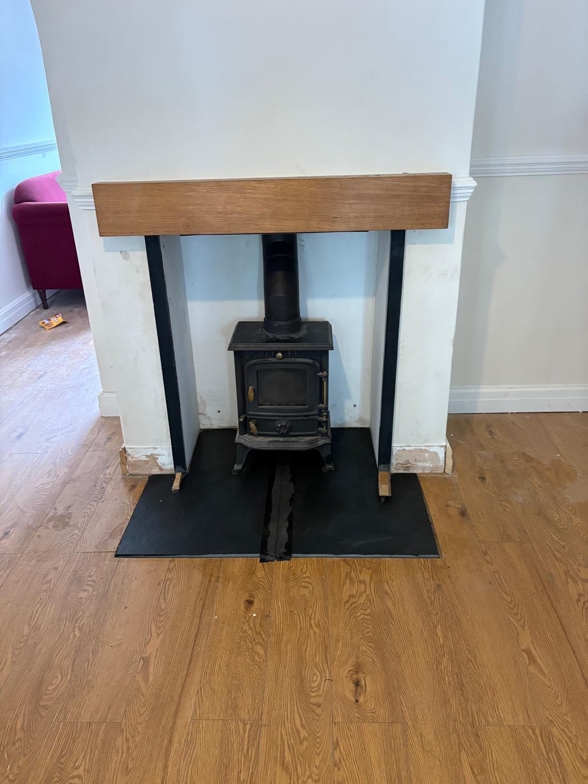 Small black cast iron stove installed in the same opening under the oak beam, on a black hearth over oak flooring
