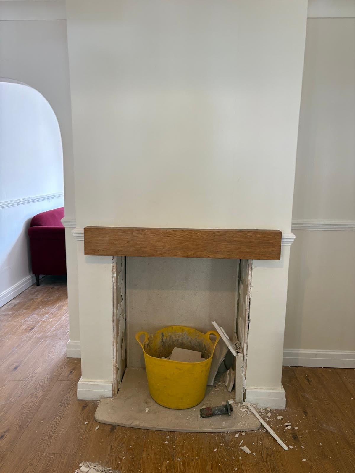 Bare plastered fireplace opening beneath an oak beam mantel, with a builder's bucket and tools on the floor