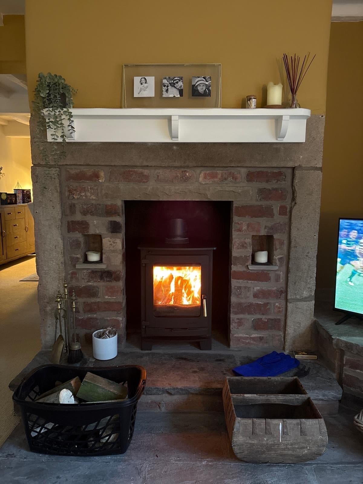 Stove alight within a stone fireplace surround, painted mantel above with candles and framed prints