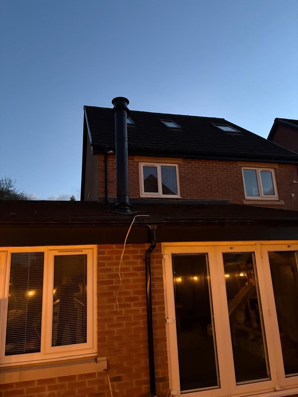 Black insulated twin-wall flue running up the exterior gable of a brick house at dusk, warm light in the windows below